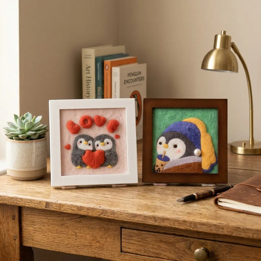 Two framed needle felted penguin pictures displayed on a wooden desk beside books and a lamp