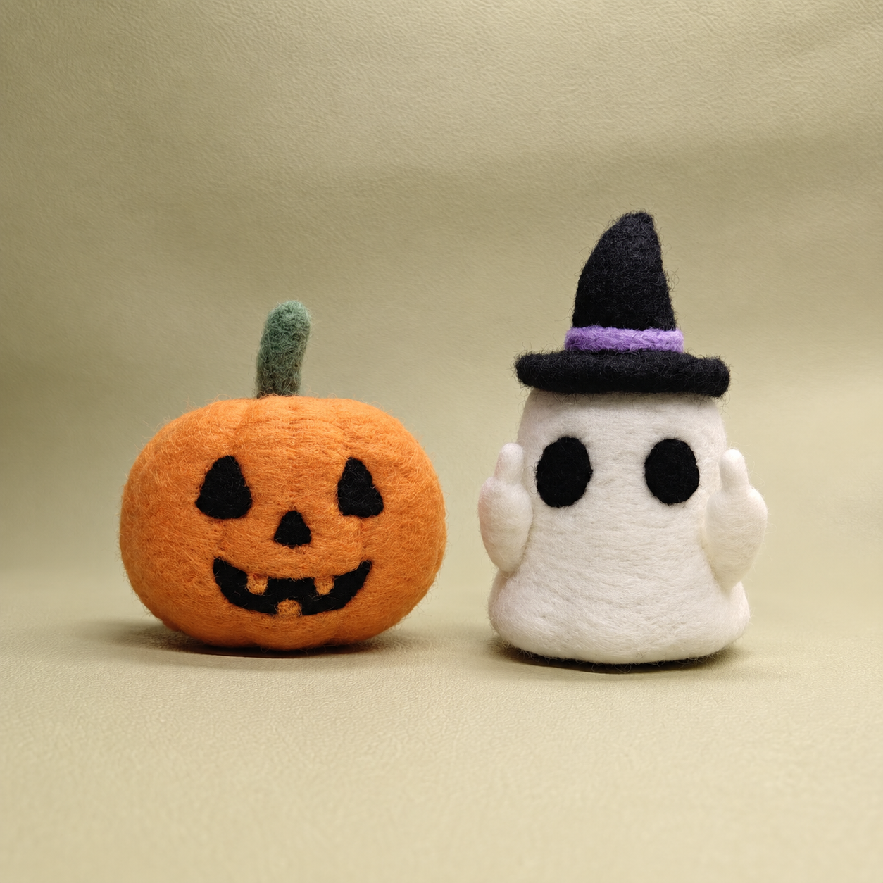 Peeka the white needle felted ghost in a black witch hat beside Gourdon the orange needle felted pumpkin on a warm beige background.